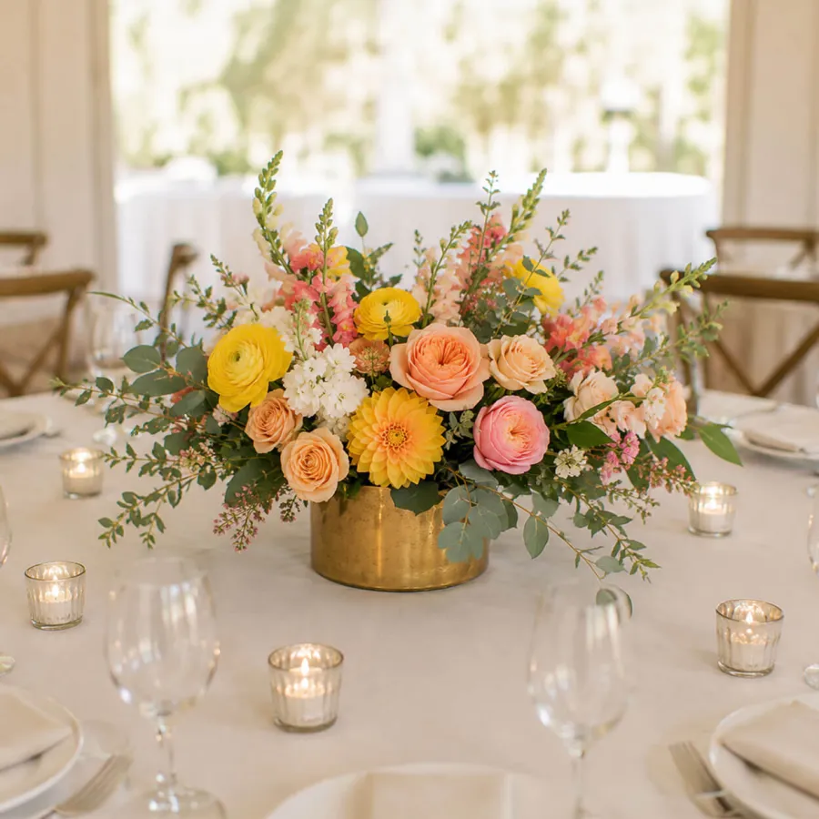 Large lush centerpiece in a gold vessel with bright yellow roses, peach ranunculus, and colorful cheerful blooms on a wedding table