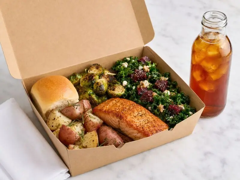Fremont Catering Company boxed dinner with seasoned salmon, roasted potatoes, Brussels sprouts, kale salad, a dinner roll, and iced tea.