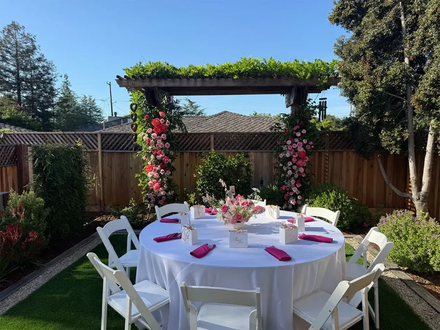 Blush-to-crimson rose arch arcing over a garden round dressed in white cloth with fuchsia napkins — bespoke floral installations from Fremont catering